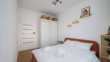 A bedroom with a bed, white walls, a wardrobe, a ceiling light fixture, a framed picture, and a small wooden shelf.