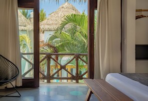 Ein Zimmer mit einem Bett, einem Sessel und Blick auf ein Gebäude mit einem Strohdach sowie Palmen.
