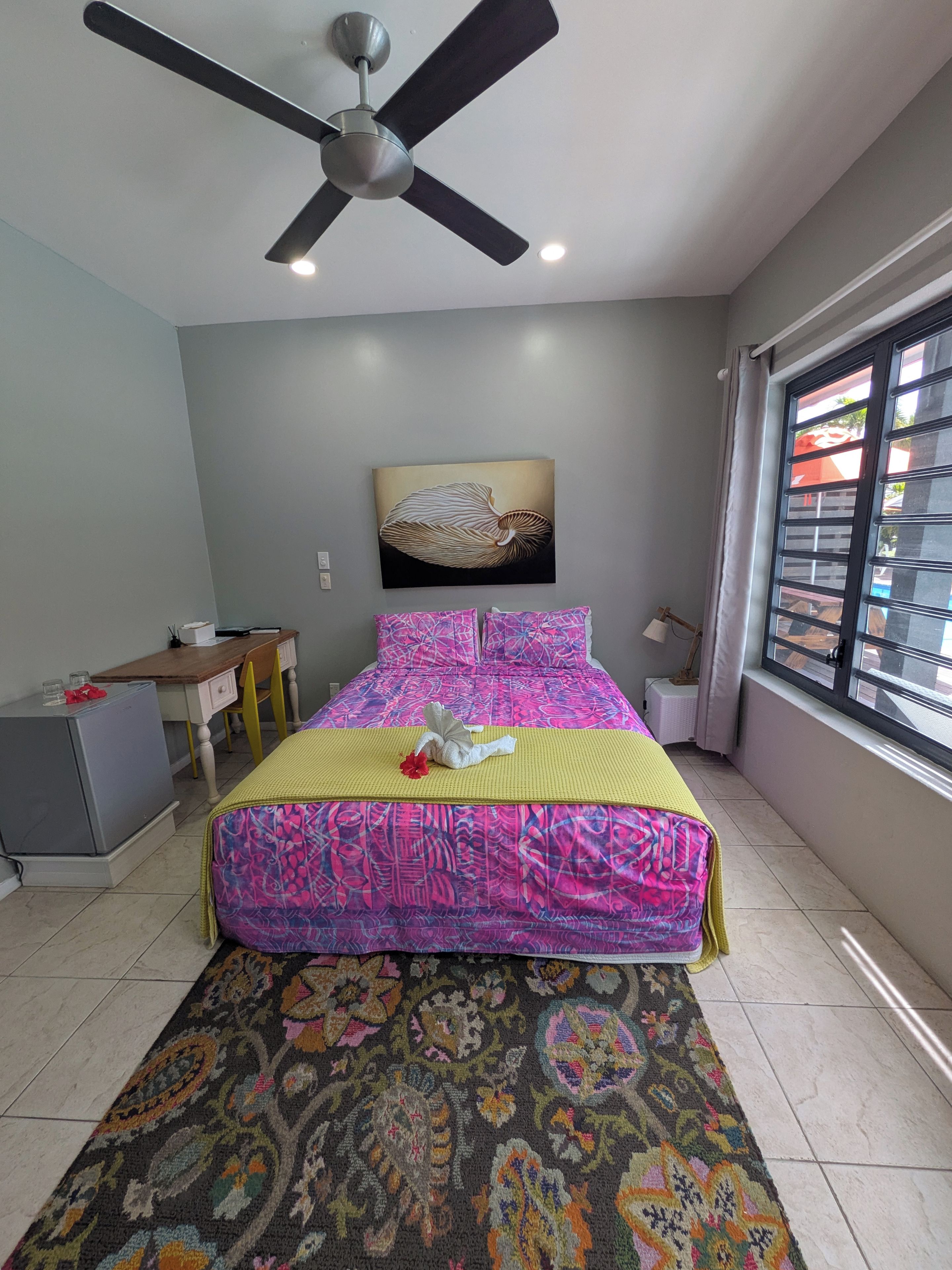 A bedroom with a bed, a desk, a ceiling fan, a window with blinds, and a wall-mounted artwork.