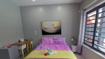 A bedroom with a bed, a desk, a ceiling fan, a window with blinds, and a wall-mounted artwork.