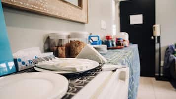 Habitación de hotel con zona de comedor que incluye una mesa con platos, tazones y una cafetera. Se ven recipientes con snacks y un espejo en la pared.