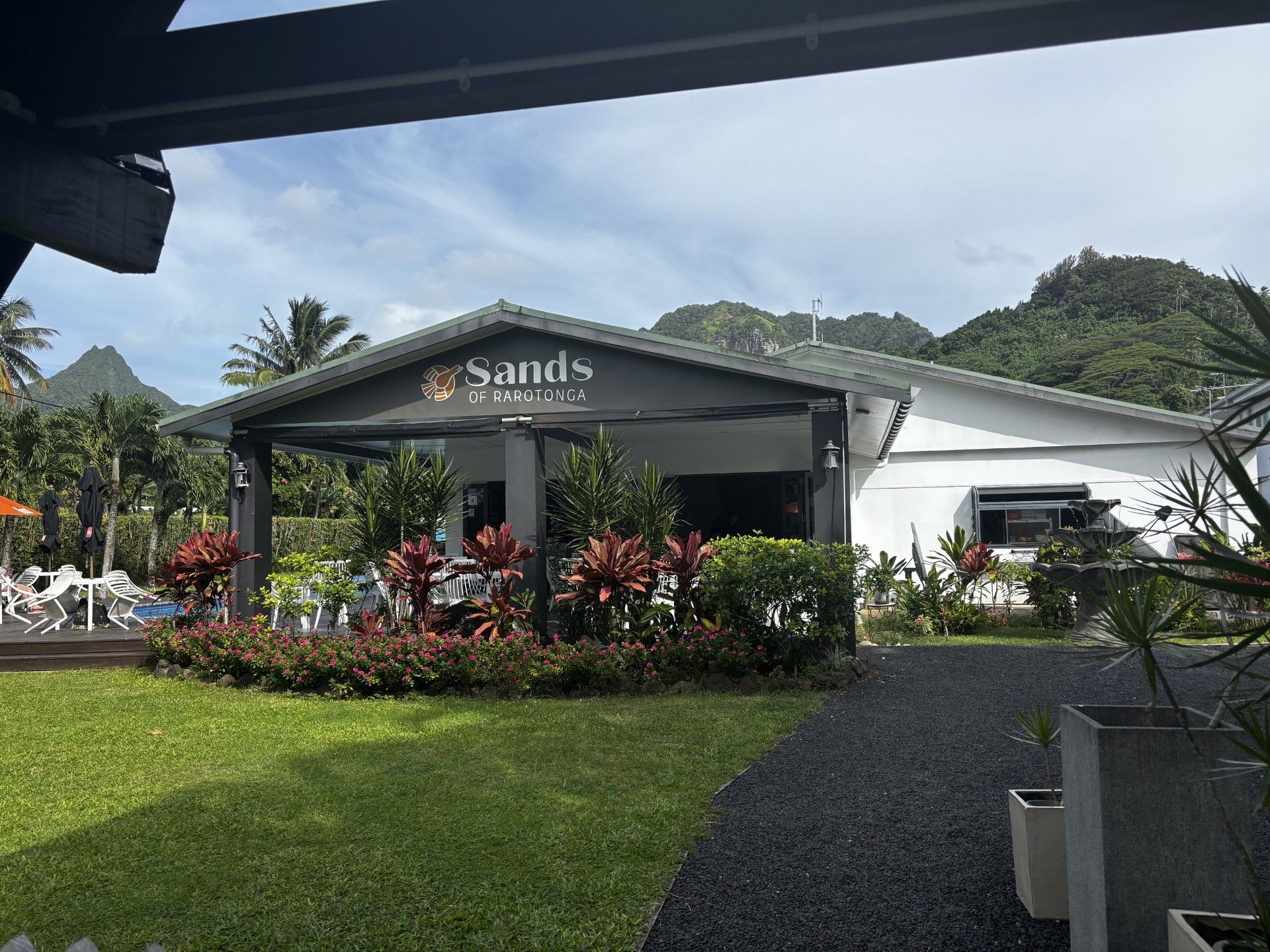 Un hotel llamado 'Sands of Rarotonga' con un espacio exterior cubierto, abundante vegetación y montañas al fondo.
