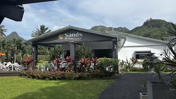 Un hotel llamado 'Sands of Rarotonga' con un espacio exterior cubierto, abundante vegetación y montañas al fondo.