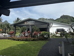 Un hotel llamado 'Sands of Rarotonga' con un espacio exterior cubierto, abundante vegetación y montañas al fondo.