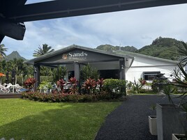 Un hotel llamado 'Sands of Rarotonga' con un espacio exterior cubierto, abundante vegetación y montañas al fondo.