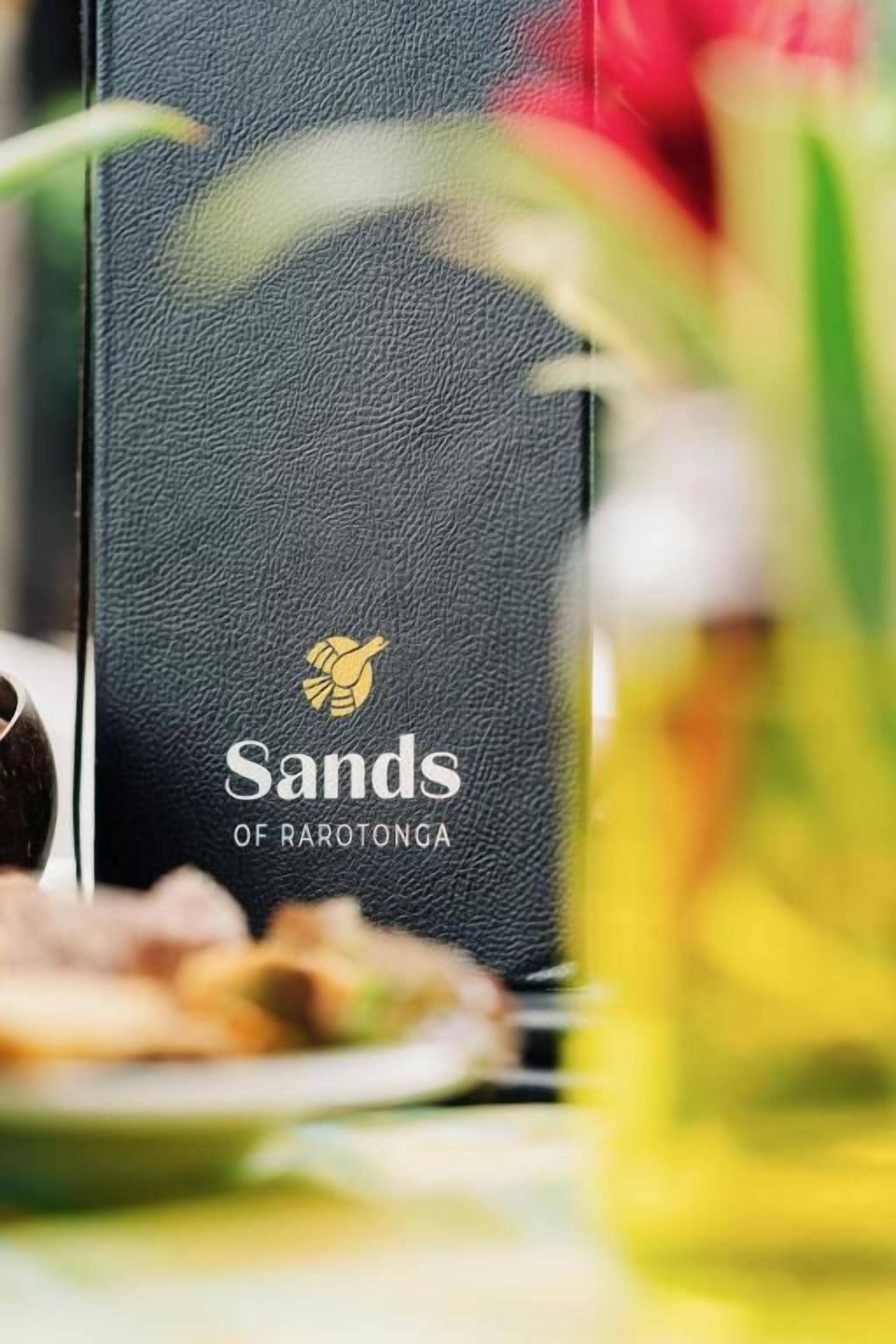 A book titled 'Sands of Rarotonga' with a logo, placed on a table with a blurred background.
