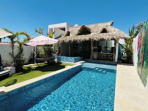 Una piscina con una estructura de techo de paja, una hamaca y un pequeño jardín.