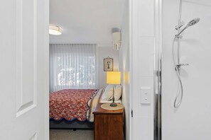 Habitación de hotel con cama, mesita de noche con lámpara, ducha y ventana con cortinas.