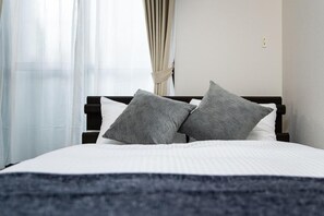 Ein ordentlich bezogenes Bett mit dunklem Kopfteil und zwei Kissen, ein Fenster mit leichtem Vorhangstoff, und eine schlichte Wand mit Lichtschalter.