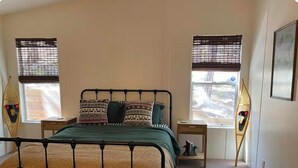Une chambre à coucher avec un lit en métal, une couvre-lit verte, des coussins à motifs et deux fenêtres équipées de stores.