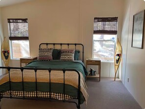 Een slaapkamer met een metalen bedframe, een groene spreidek, gestreepte kussens en twee ramen met luiken.