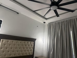 Una habitación con ventilador de techo, cabecera y ventana con cortinas.