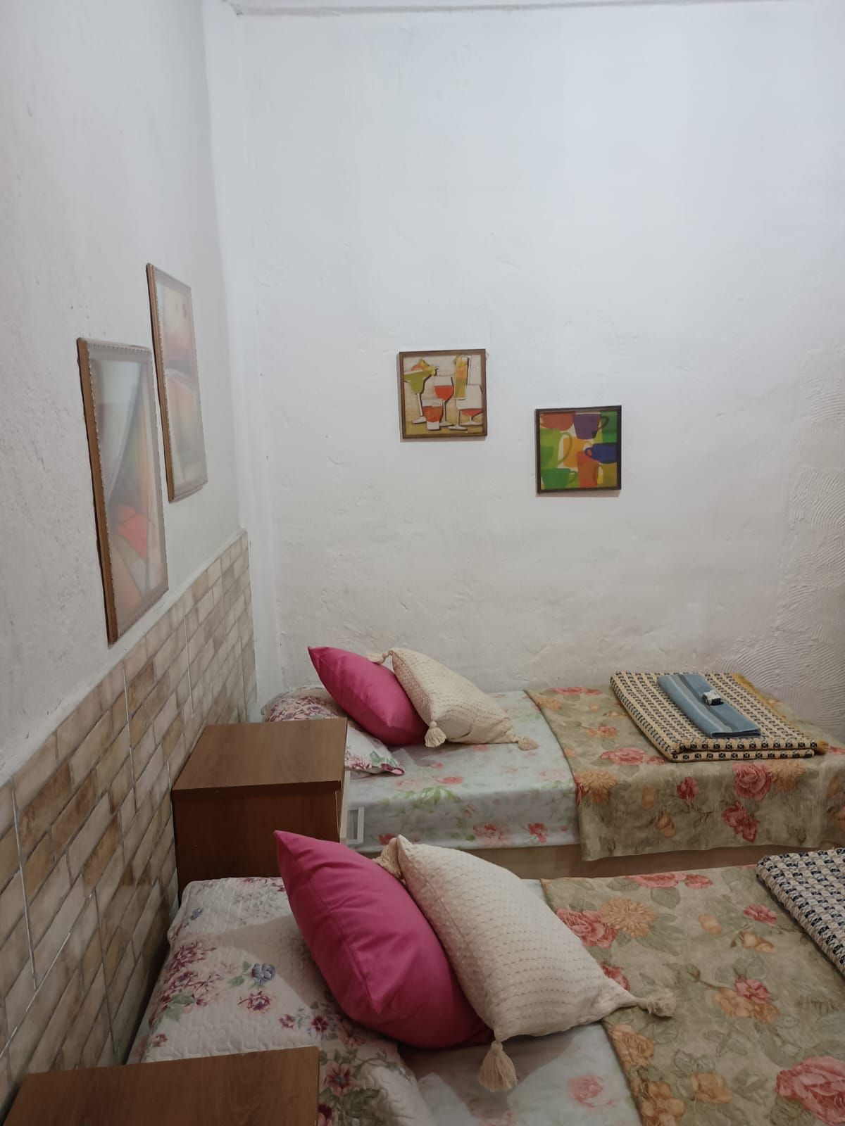 A small, simple room with two beds, each with floral bedding and pillows, a brick-patterned headboard, and two framed artworks on the wall.