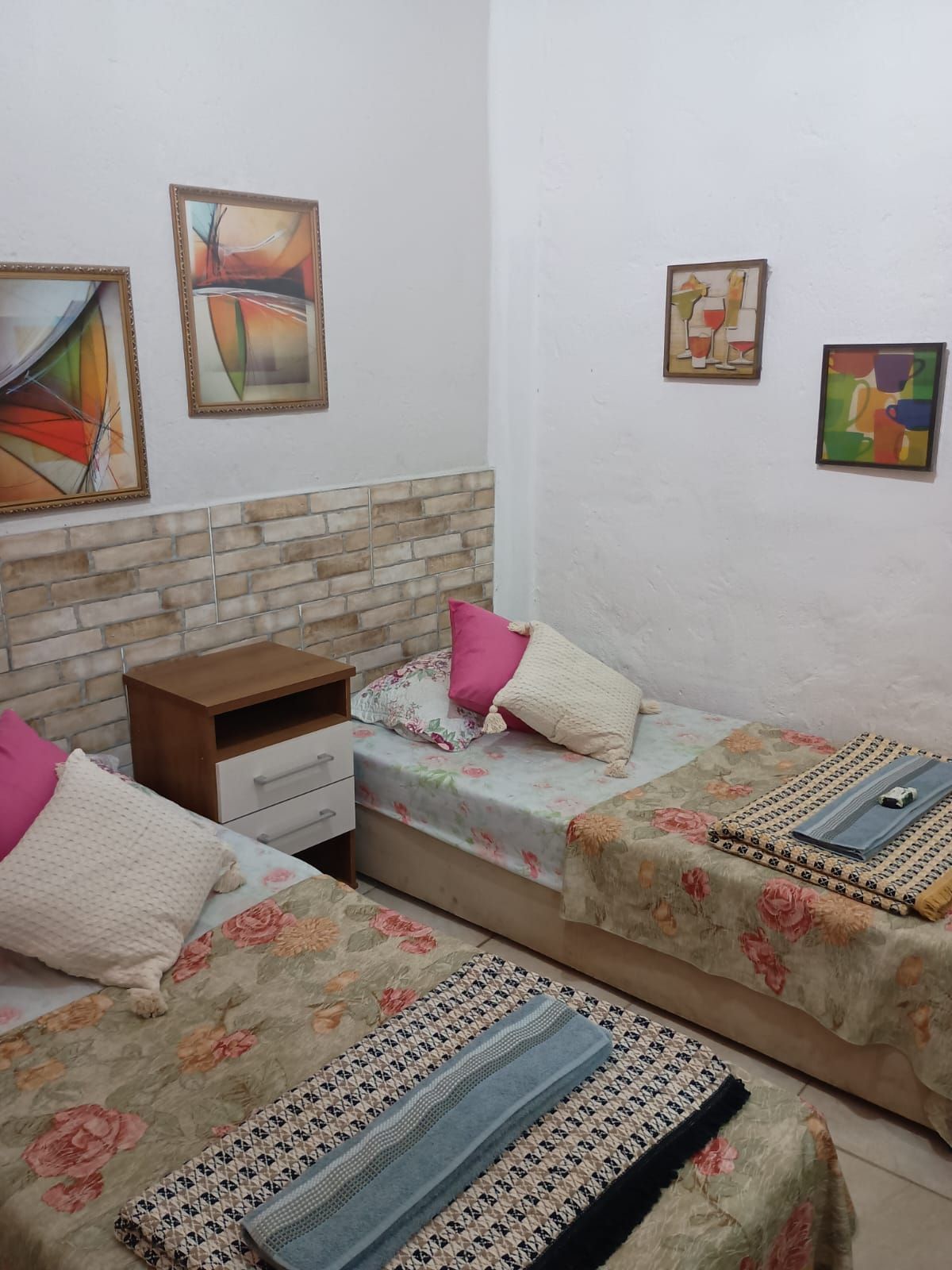 A bedroom with two beds, a nightstand, and a dresser. There are three paintings on the wall.