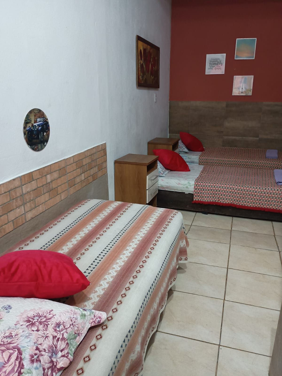 Una habitación con dos camas, cada una con cubrecama estampado y almohadas rojas, una mesita de noche de madera y una pared con un cuadro enmarcado y un espejo pequeño.