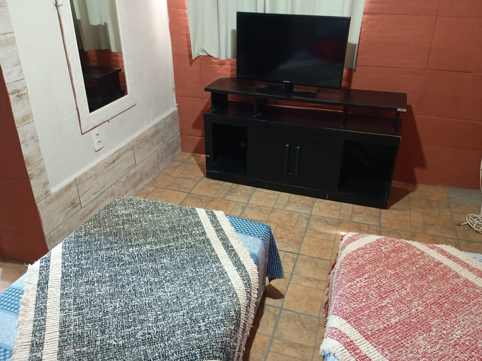 A room with a television on a black stand, a window with curtains, a tiled floor, and two beds with blankets.