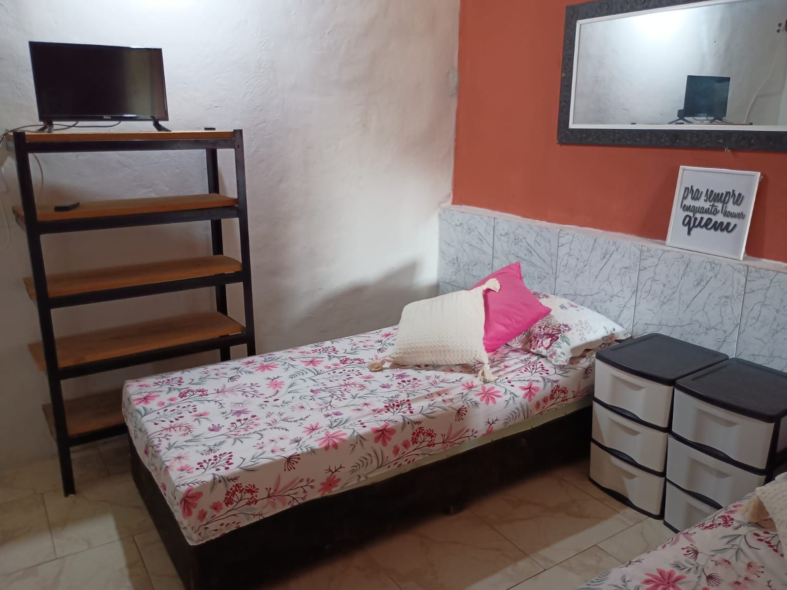 Una habitación pequeña con cama individual, una estantería de madera, un televisor encima, ropa de cama floral, una mesita de noche con cajones y un espejo en la pared.