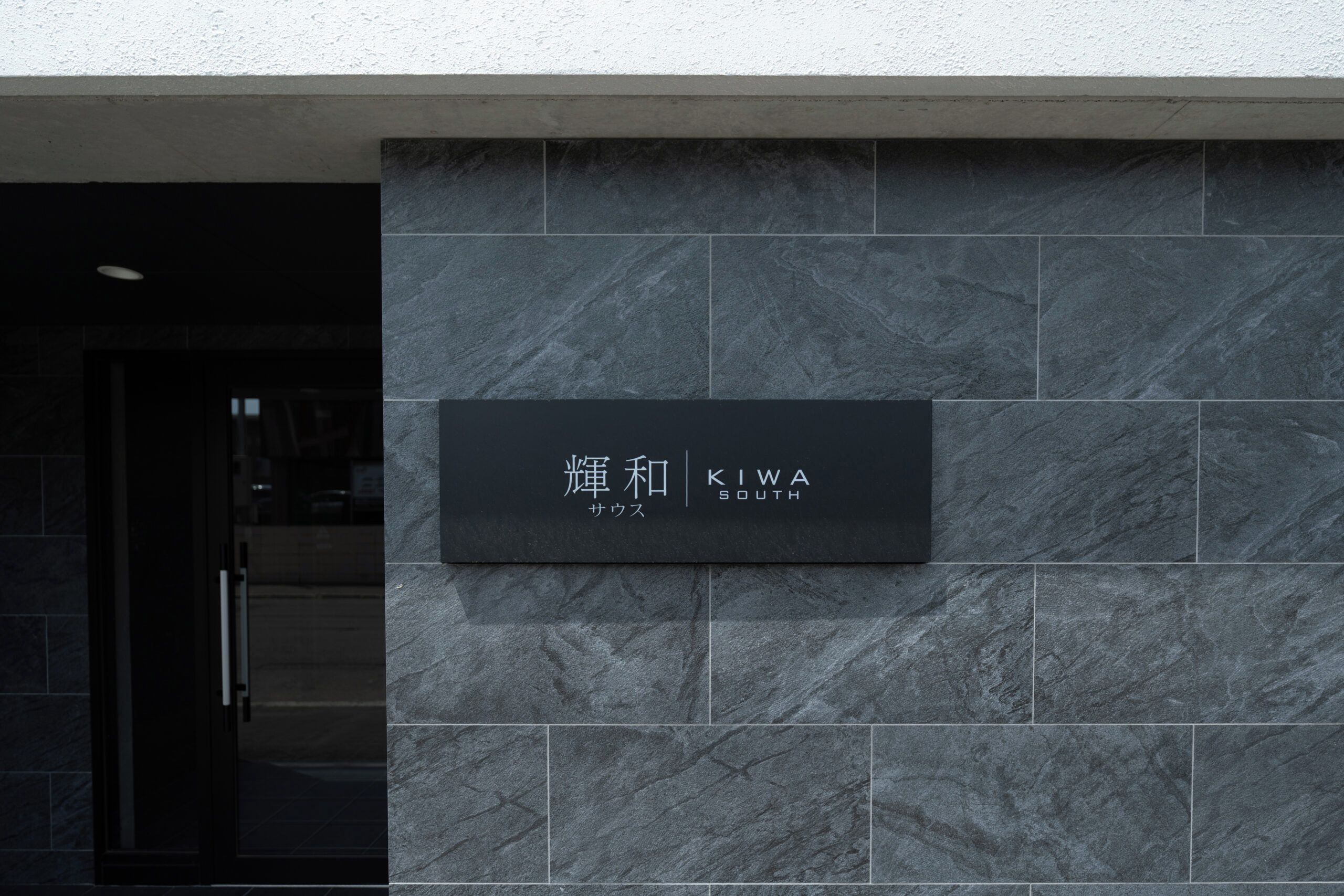 A black sign with white text on a grey stone wall, indicating the name 'KIWA SOUTH' and the word 'SAWUS'.
