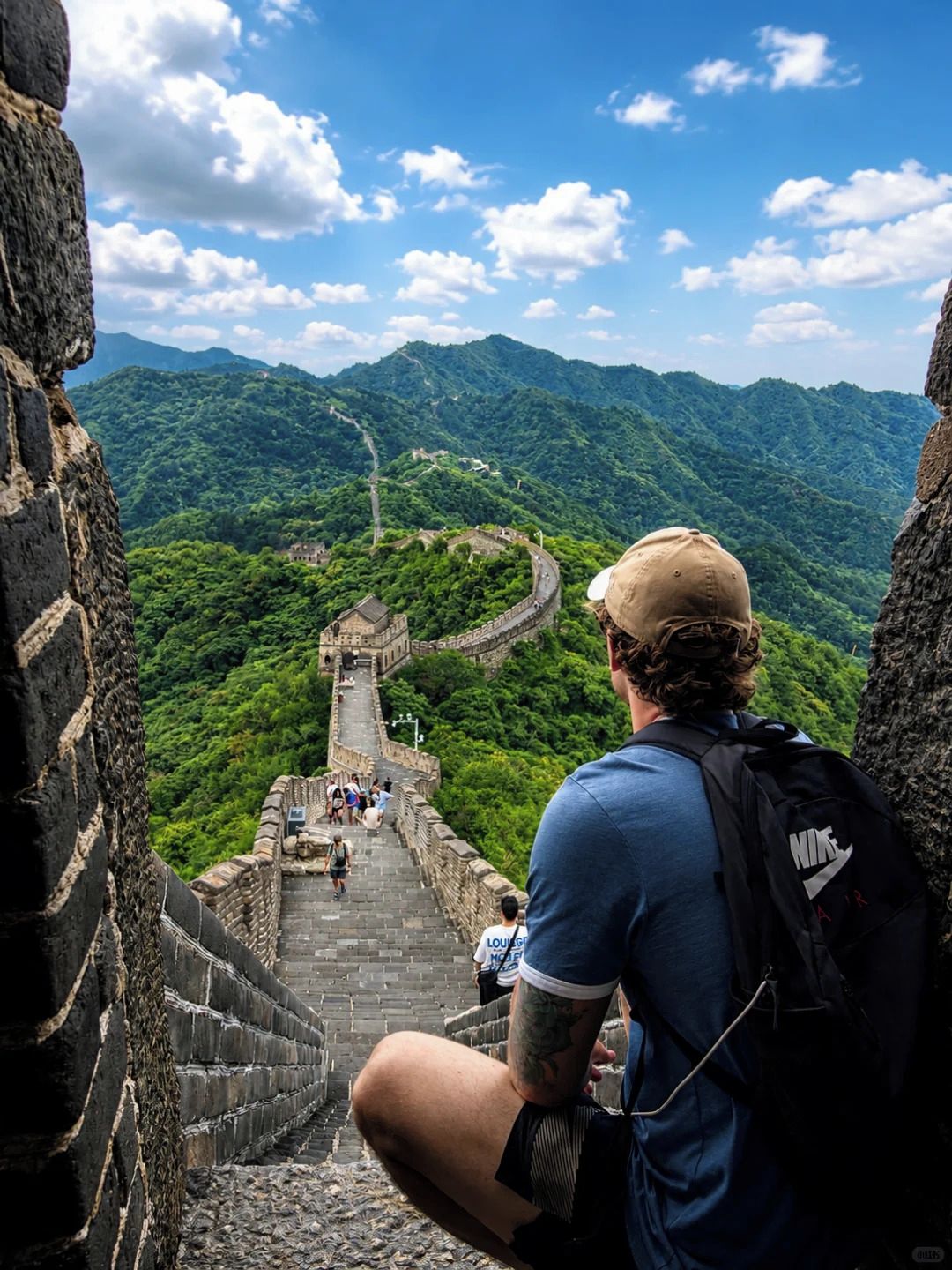 Una persona sentada sobre un muro de piedra contemplando la Gran Muralla China, con exuberantes montañas verdes al fondo.
