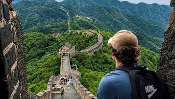 A person sitting on a stone wall overlooking the Great Wall of China with lush green mountains in the background.