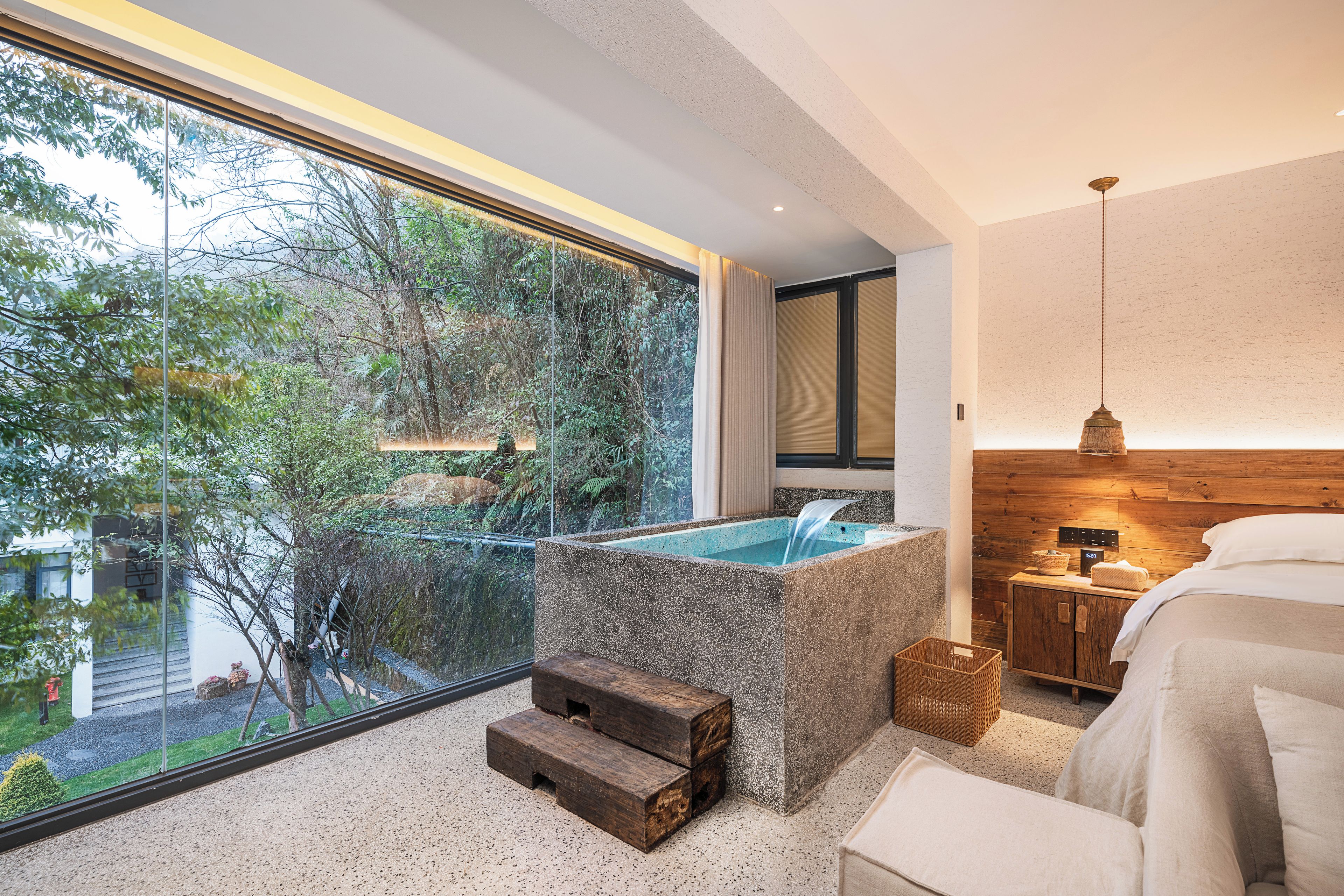 A modern bathroom with a freestanding bathtub, a wooden bench, and a view of a lush garden through a large window.