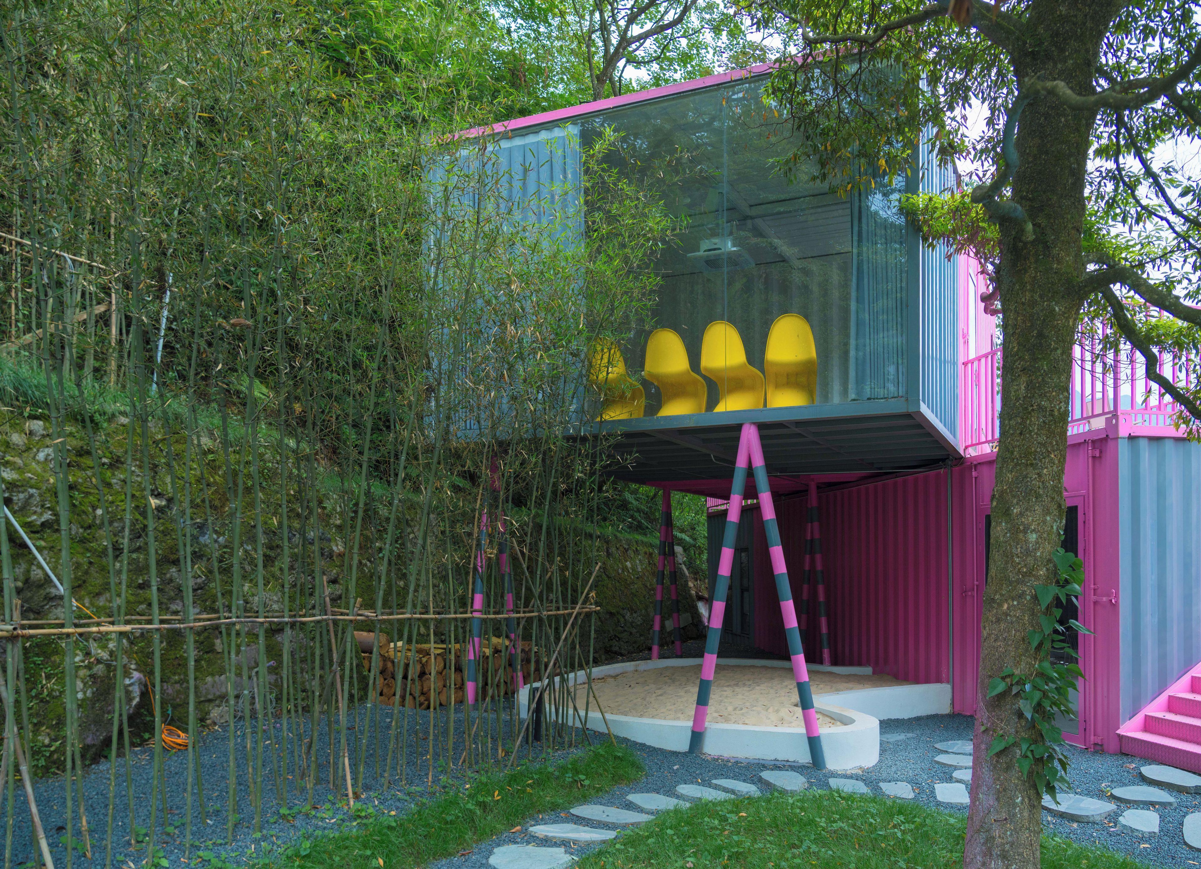 Una estructura moderna al aire libre con cerramiento de vidrio, terraza de madera y un edificio de color rosa y azul.