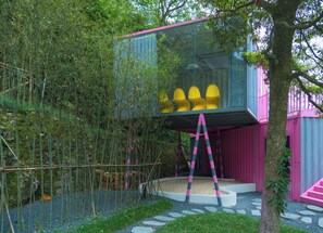 Una estructura moderna al aire libre con cerramiento de vidrio, terraza de madera y un edificio de color rosa y azul.