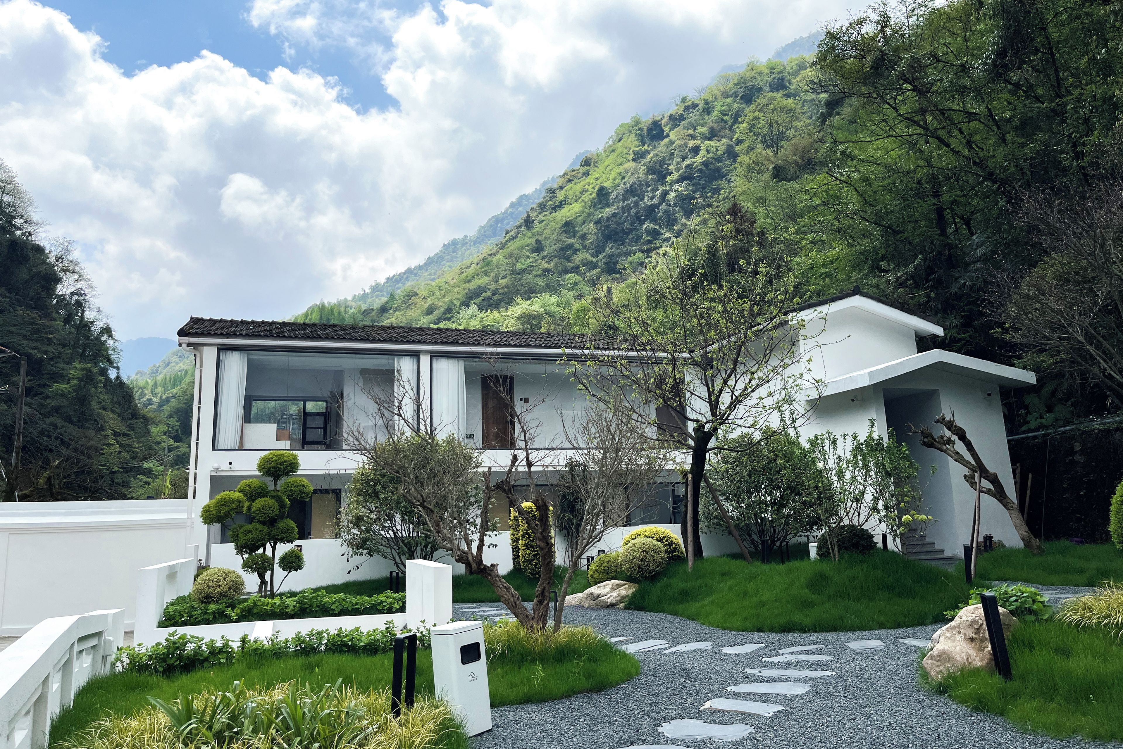 A modern two-story house with a white exterior, surrounded by lush greenery and a mountainous backdrop.