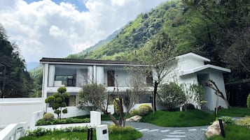 A modern two-story house with a white exterior, surrounded by lush greenery and a mountainous backdrop.