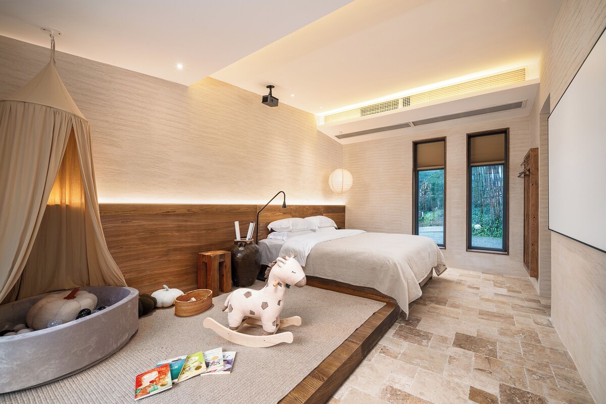 A modern bedroom with a large bed, a wooden headboard, a small seating area with a rocking horse, and a view of the outdoors through large windows.