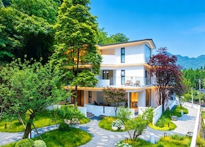 A multi-story building with a balcony, surrounded by lush greenery and a clear sky.