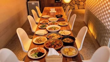 Una larga mesa de madera con varios platos de comida, sillas blancas y una pared de piedra con luces colgantes.