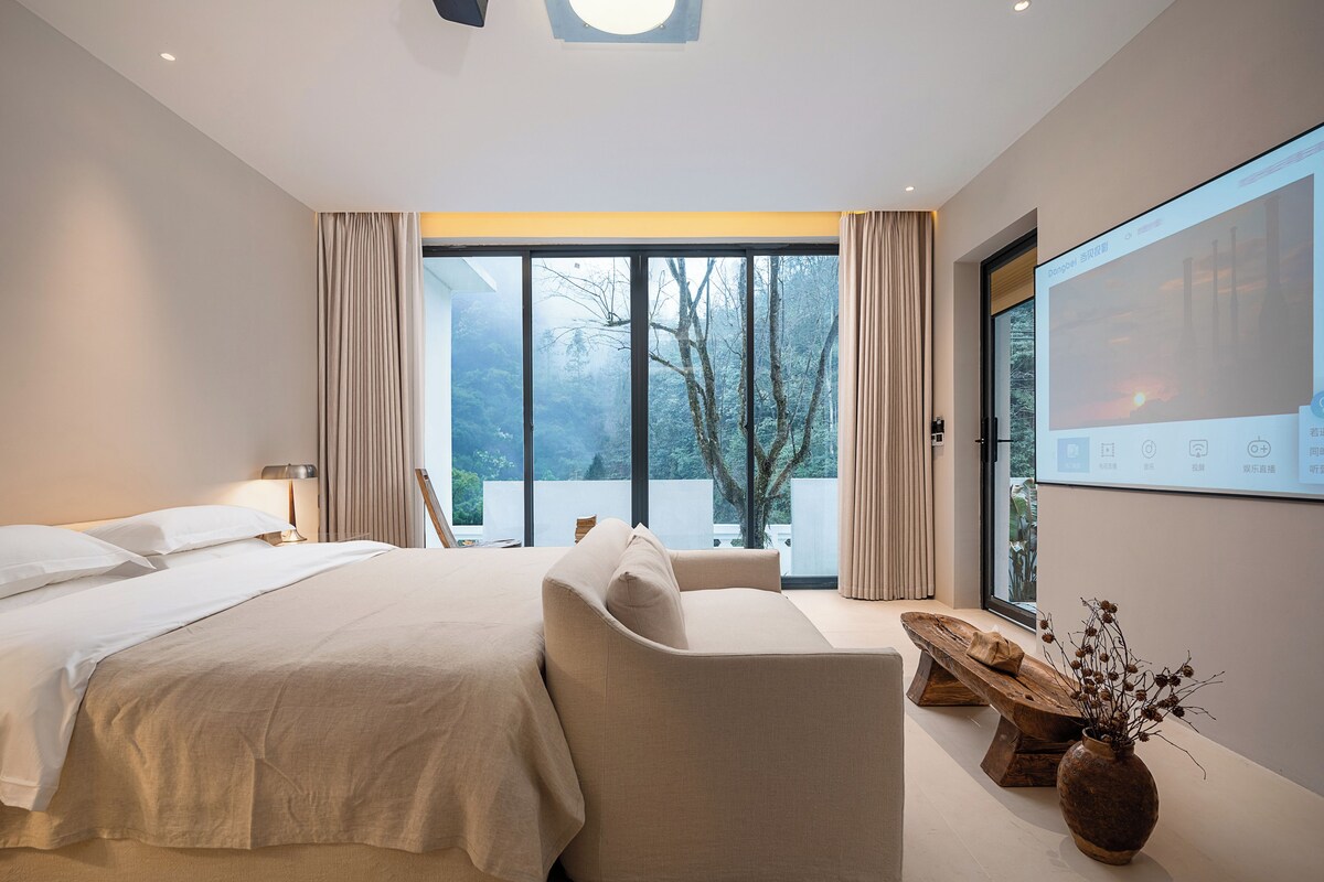 A modern bedroom with a large bed, a sofa, a wooden side table, and a large window with a view of trees.