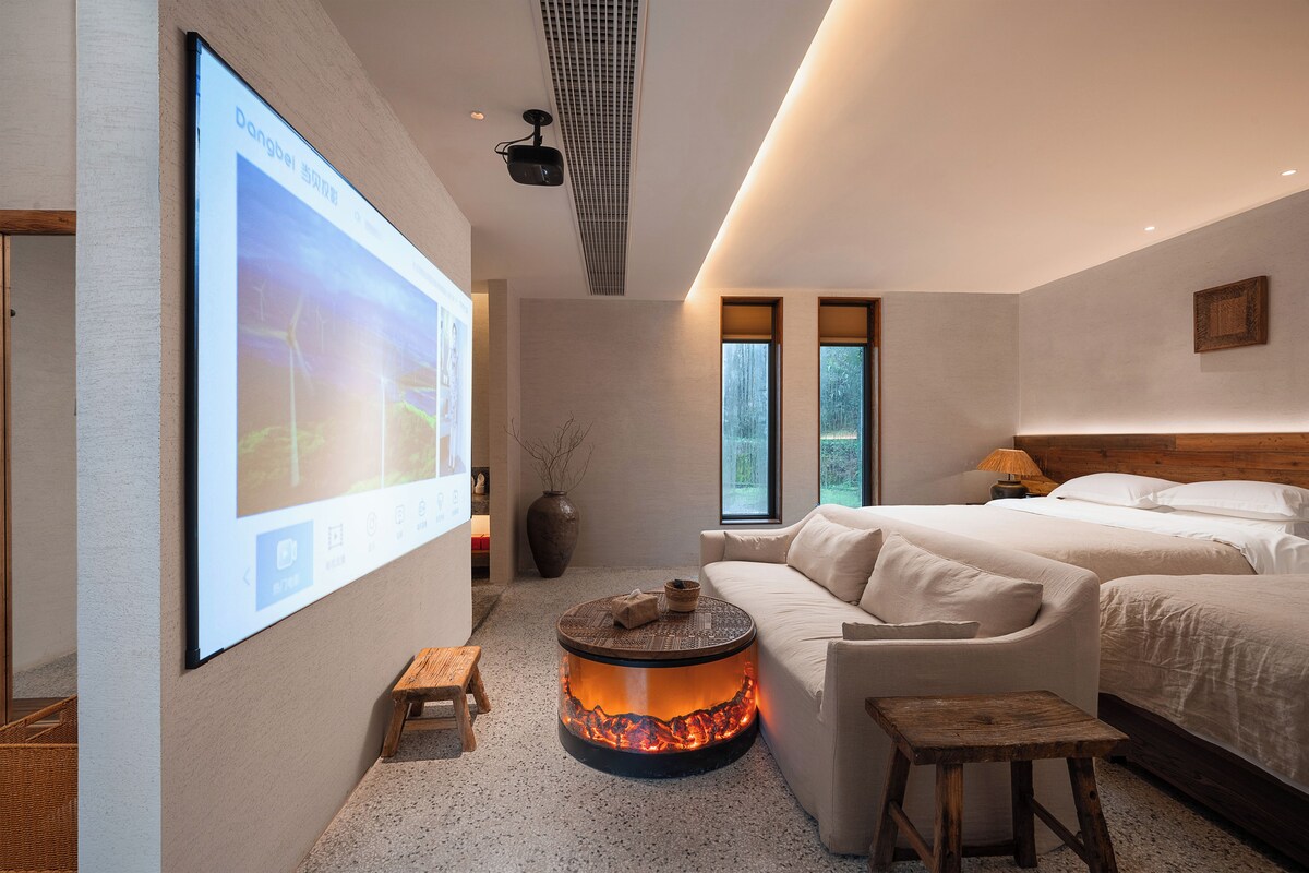 A modern hotel room with a large screen, a fireplace, a sofa, and two beds.