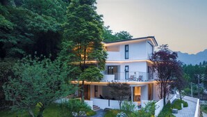 Uma casa moderna de dois andares com uma varanda, cercada por um jardim bem cuidado e árvores.