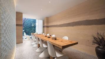 Un comedor moderno con una larga mesa de madera, sillas blancas y vista a árboles a través de amplios ventanales.