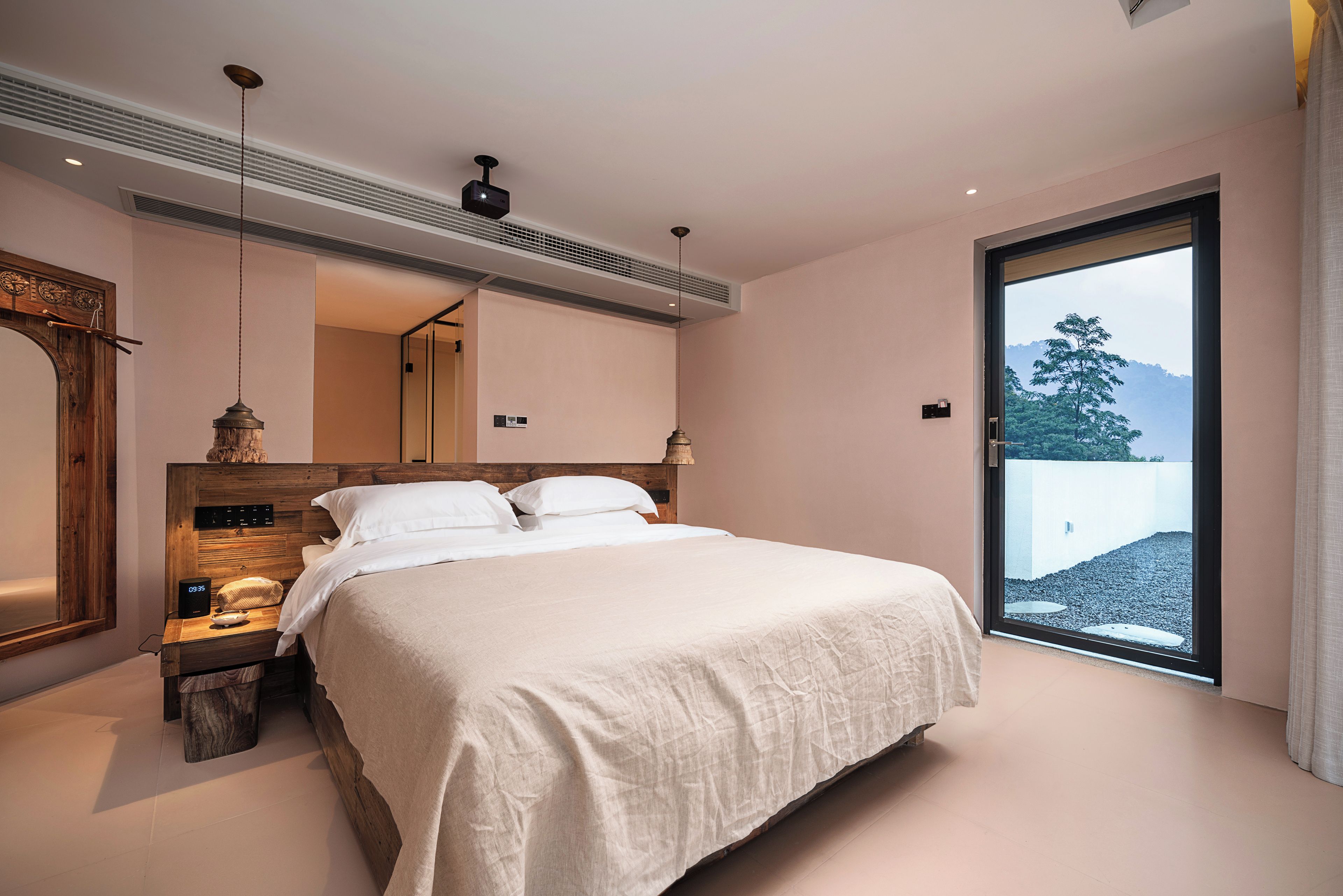 Un dormitorio moderno con una cama grande, cabecera de madera, mesita de noche, un reloj en la pared y una ventana con vista a árboles.