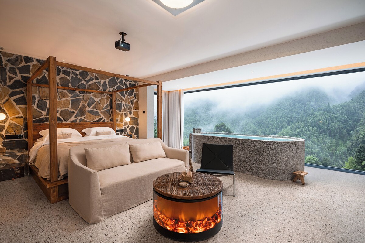 A bedroom with a stone wall, a bed, a sofa, a fireplace, and a view of a forest.