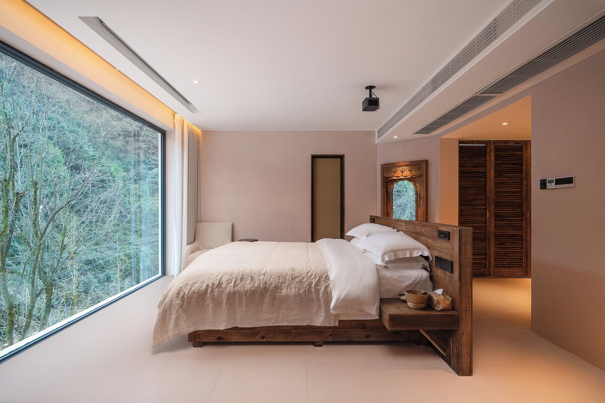 A modern bedroom with a large window, a wooden bed frame, and a view of trees.