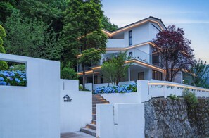 Un bâtiment moderne à plusieurs étages, doté d’un escalier menant à l’entrée, et entouré d’une végétation luxuriante et de buissons d’hortensias bleus.