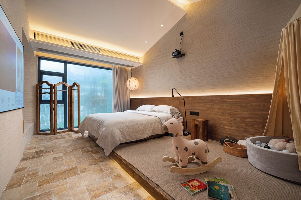 A modern bedroom with a large bed, a rocking chair, a side table, and a view of the outdoors.