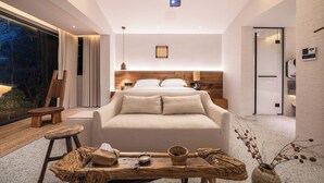 Quarto moderno com uma cama grande, mesas de cabeceira, cabeceira de madeira, um criado-mudo, uma pequena mesa com uma tigela e uma vela, e vista do exterior pela janela.