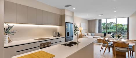 A modern kitchen with a central island, a dining area with a round table, and a view of greenery outside.