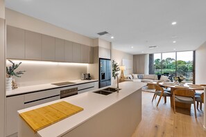 A modern kitchen with a central island, a dining area with a round table, and a view of greenery outside. - Spacious 3-Bed Townhouse In Mile End (Mile End)