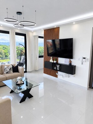 Ein modernes Wohnzimmer mit einem Glastisch, einer Couch, einem Wand-Fernseher und Blick auf Grünflächen.
