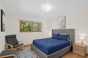 Ein Schlafzimmer mit Bett, Stuhl, Nachttisch, Lampe und Fenster mit Blick auf Bäume.
