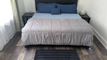 A neatly made bed with a grey duvet and pillows, a black headboard, a bedside table with a tissue box, and a dark grey rug on a wooden floor.