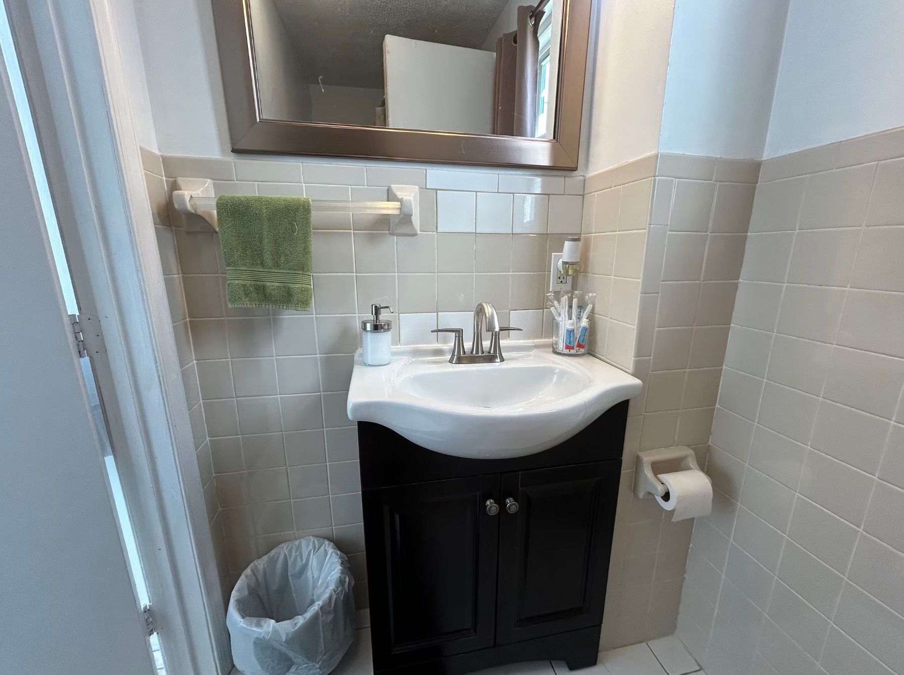 A bathroom with a white sink, a mirror, a towel rack with a green towel, a trash bin, and a toilet paper holder.