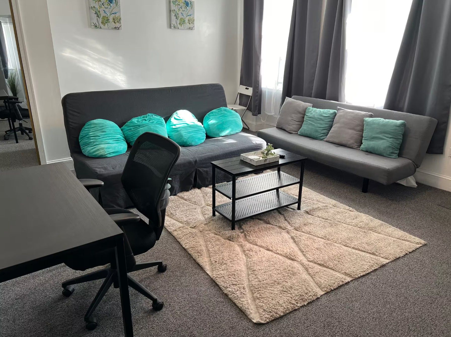 A modern living room with a black office chair, a grey sofa with teal cushions, a coffee table, and a patterned rug.