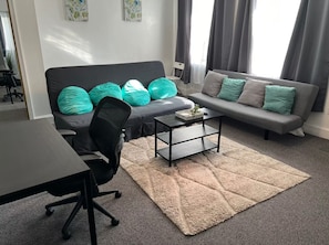 A modern living room with a black office chair, a grey sofa with teal cushions, a coffee table, and a patterned rug.